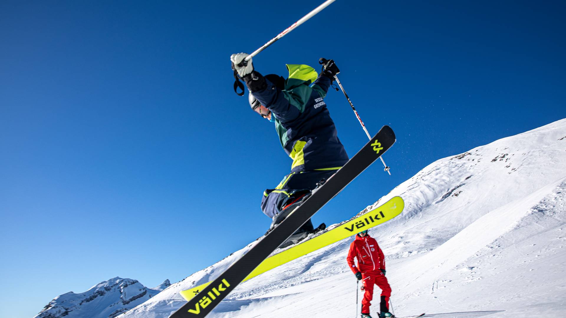 Skier jumping in the air with yellow skis, helmet, and poles; guide in red on snow-covered mountain.
